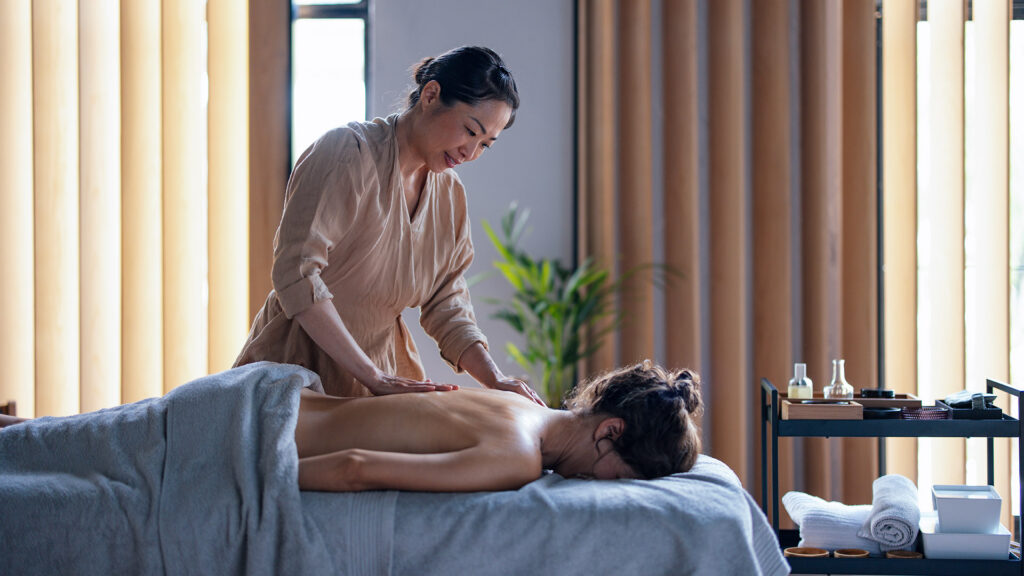 Beautiful Asian female masseuse giving a massage to a woman lying face down on the bed. They are in a spa center or a massage parlour.