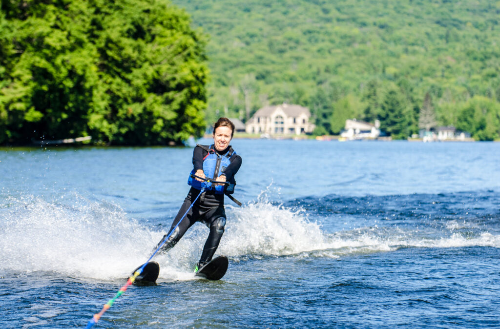 Woman in wet suit water skiing and smiling, in a lake, with chalets in background during a summer day