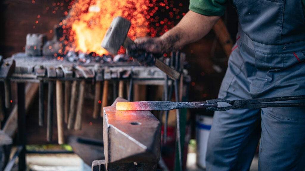 An unrecognizable blacksmith forges hot metal with a hammer on an anvile.