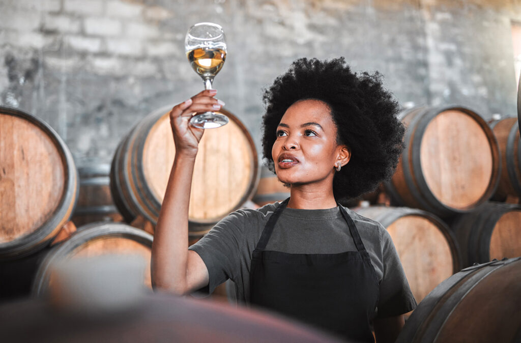 Black woman tasting wine at a winery, looking and checking the color and quality of the years produce. Young African American sommelier proud of the new addition, analyzing white wine in a cellar