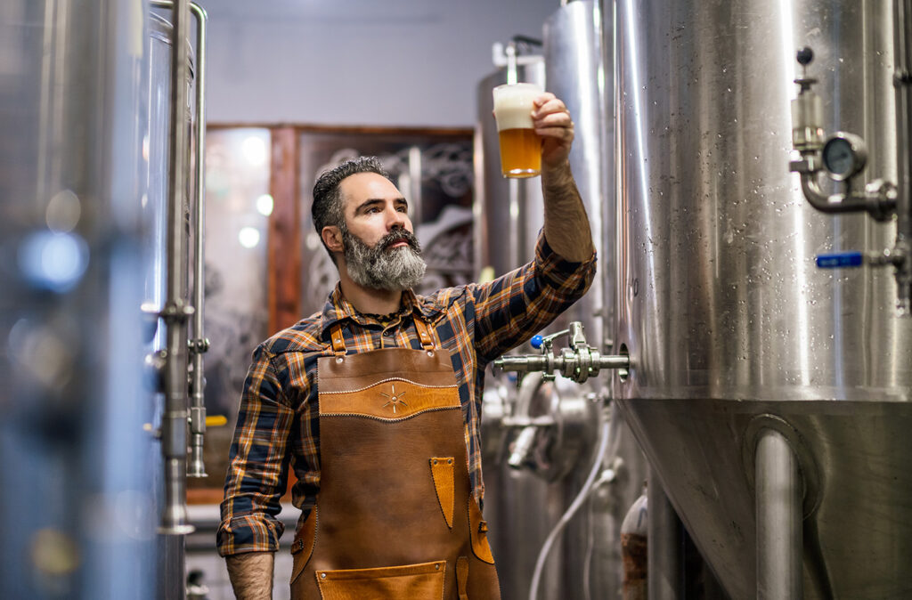 Bearded brewery master holding glass of beer and evaluating its visual characteristics. Small family business, production of craft beer.