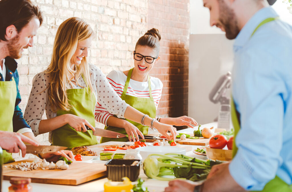 Group of people wearing aprons taking part in cooking class, preparing food, slicing vegetables, talking, laughing.