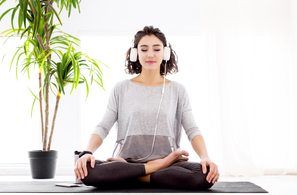 Slender female folded crossed legs in lotus position. Young athletic woman in fitness tracker and headphones is practicing yoga, meditating, relaxing while sitting in padma asana at home.