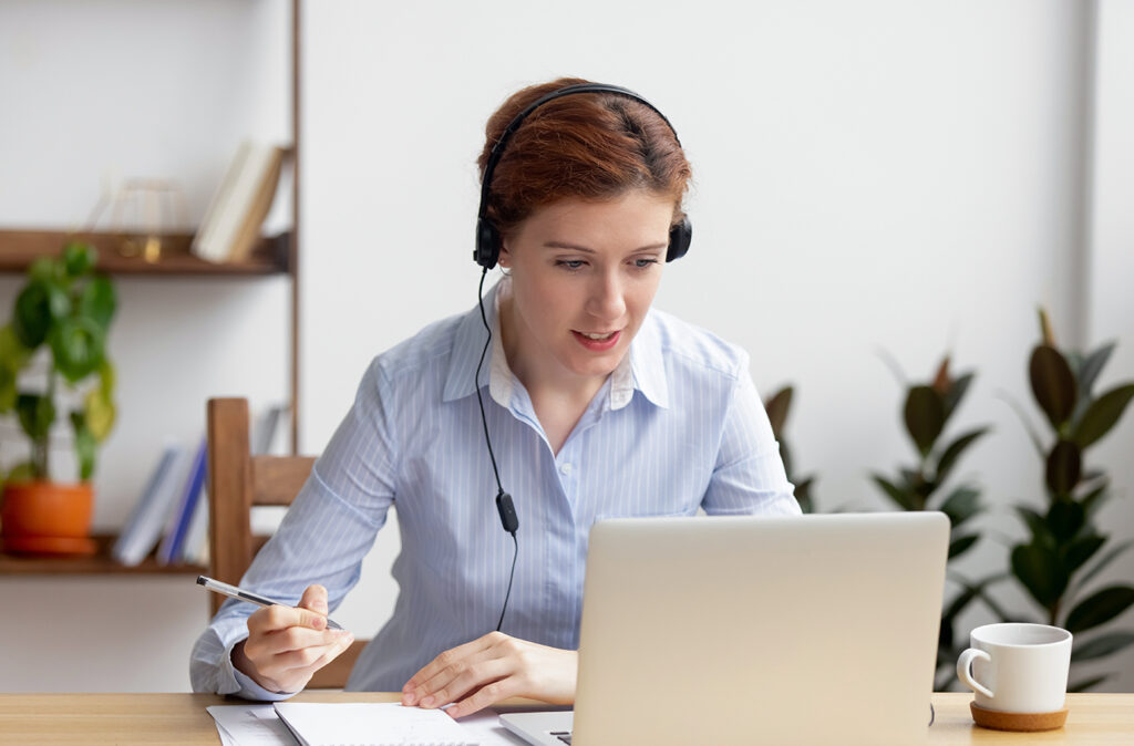 Businesswoman wearing headphones watching video webinar making conference online call writing notes talking on skype, focused woman study online looking at laptop listening translating lecture course