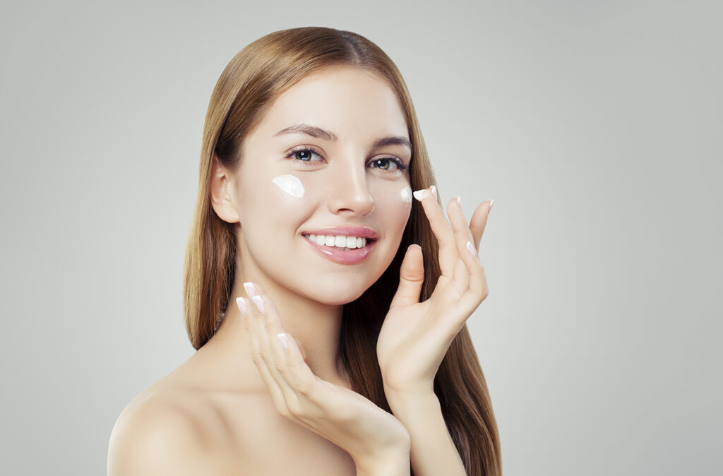 Happy young woman applying cream on her face. Skin care, beauty and facial treatment concept