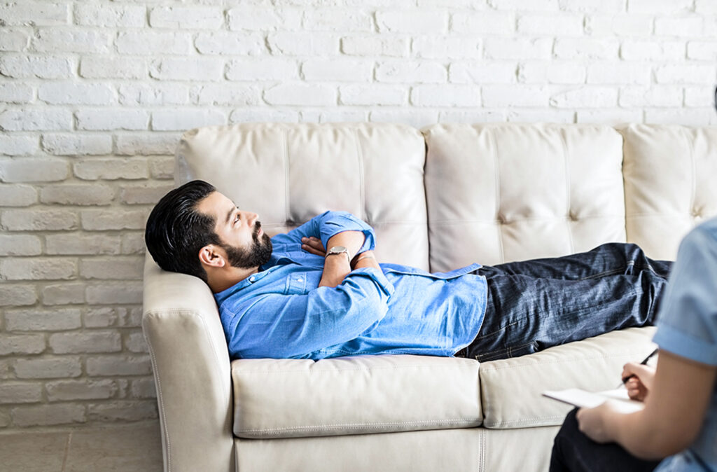 Young hispanic man lying on couch while female psychologist making notes