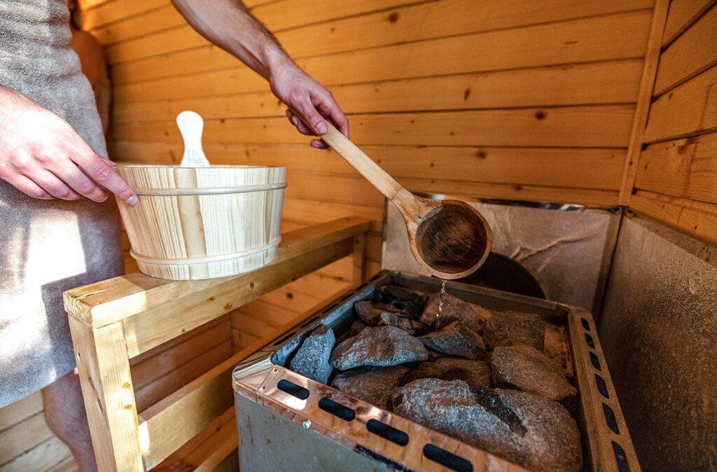 Close-up shot of unrecognizable man pouring water from wooden barrel on hot rocks in Finnish sauna.