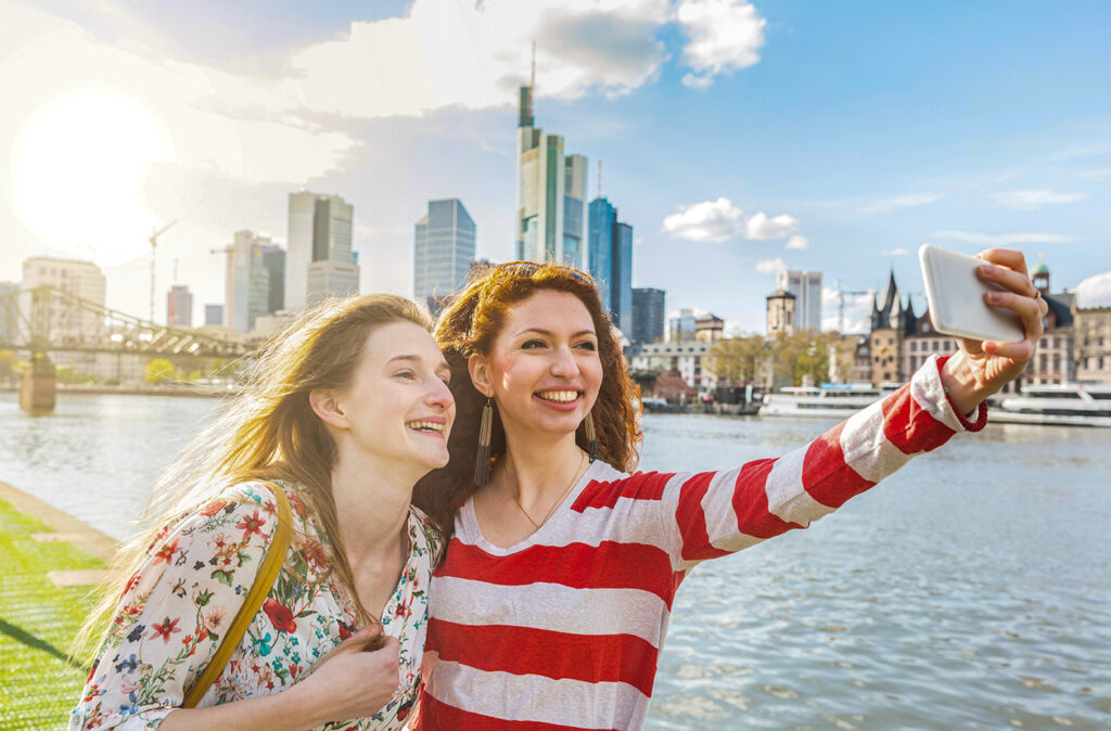 Two beautiful young women taking a selfie with Frankfurt skyline on background on a spring day. They are smiling and looking at the smart phone. Friendship and travel concepts.