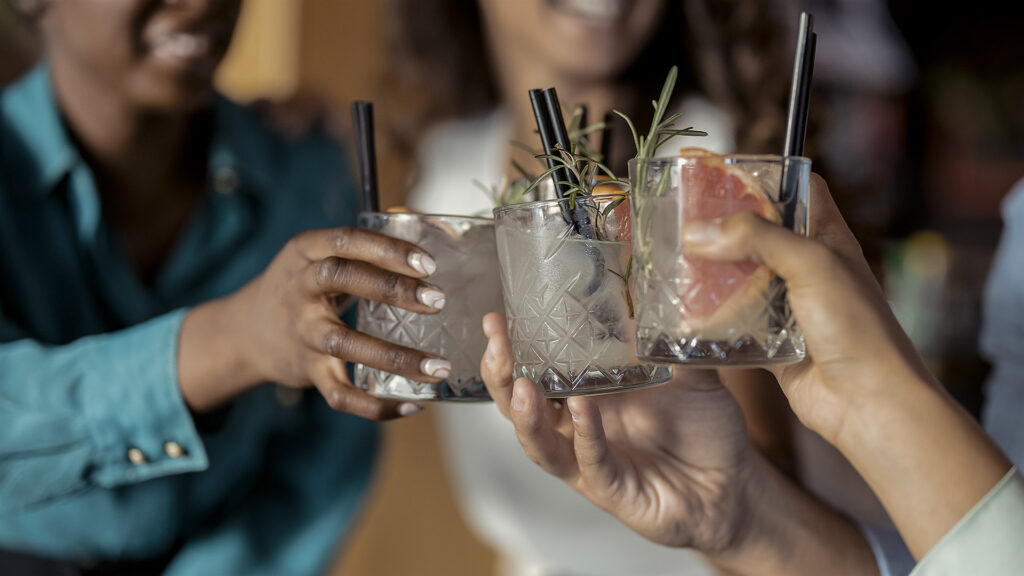 People with alcohol drinks clinking glasses in a restaurant having fun together, laughing, drinking cocktails. Party, celebration, nightlife and people concept - smiling friends clinking glasses