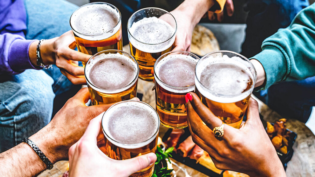 Multiracial group of young people cheering glass of beer together at brewery pub- Happy friends enjoying summer drinking blonde pint sitting at bar table- Food and beverage concept-Youth culture