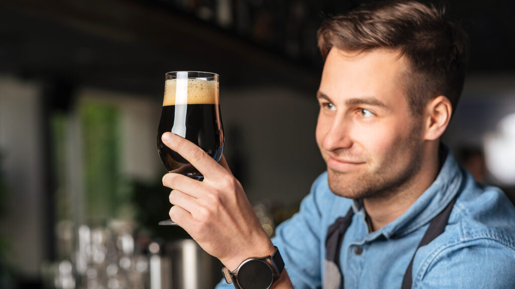 Bartender at work. Handsome man looks at glass of dark craft beer with foam in interior of bar, close up, free space