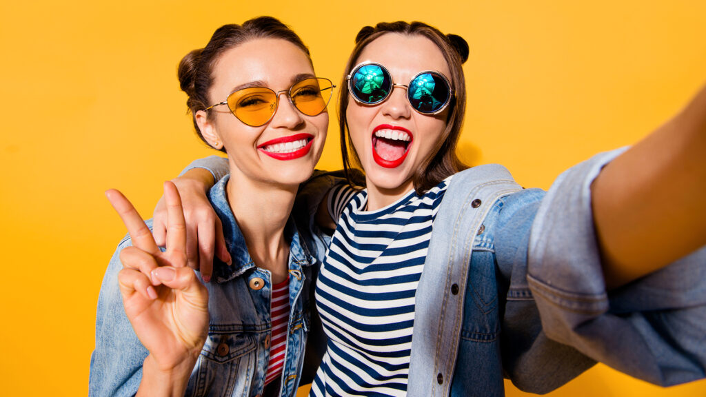 Two glad positive grinning lady stand in glasses spectacles street style stylish trendy cool casual denim jeans clothes isolated on yellow background in take picture on cellular make hollywood smile