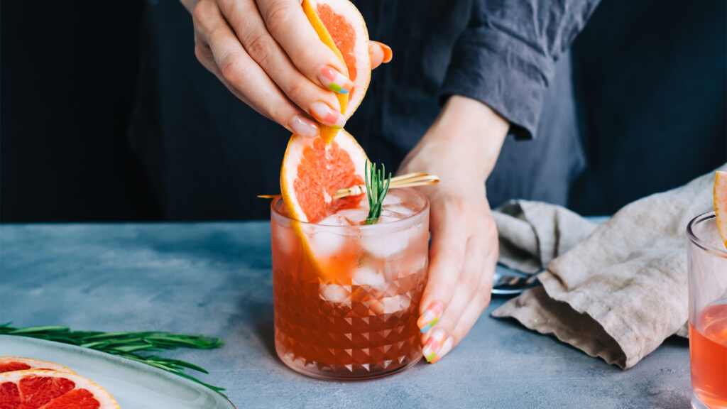 Female bartender hand squeezes juice from fresh grapefruitÂ in cocktail lemonade with ice and rosemary.