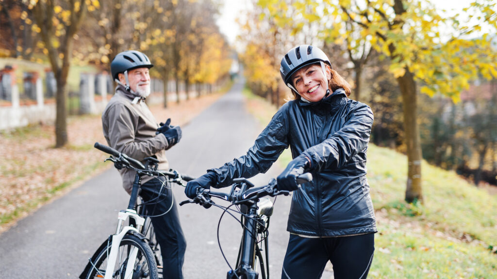 An active senior couple with helmets and electrobikes standing outdoors on a road in nature.