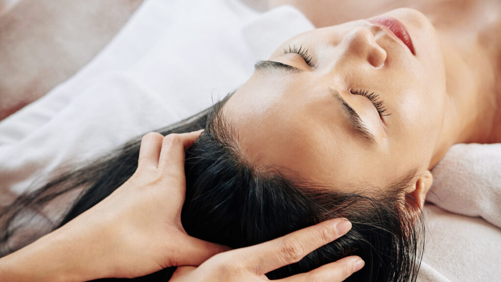 Close-up image of beautician massaging head of female client relaxing on massage bed