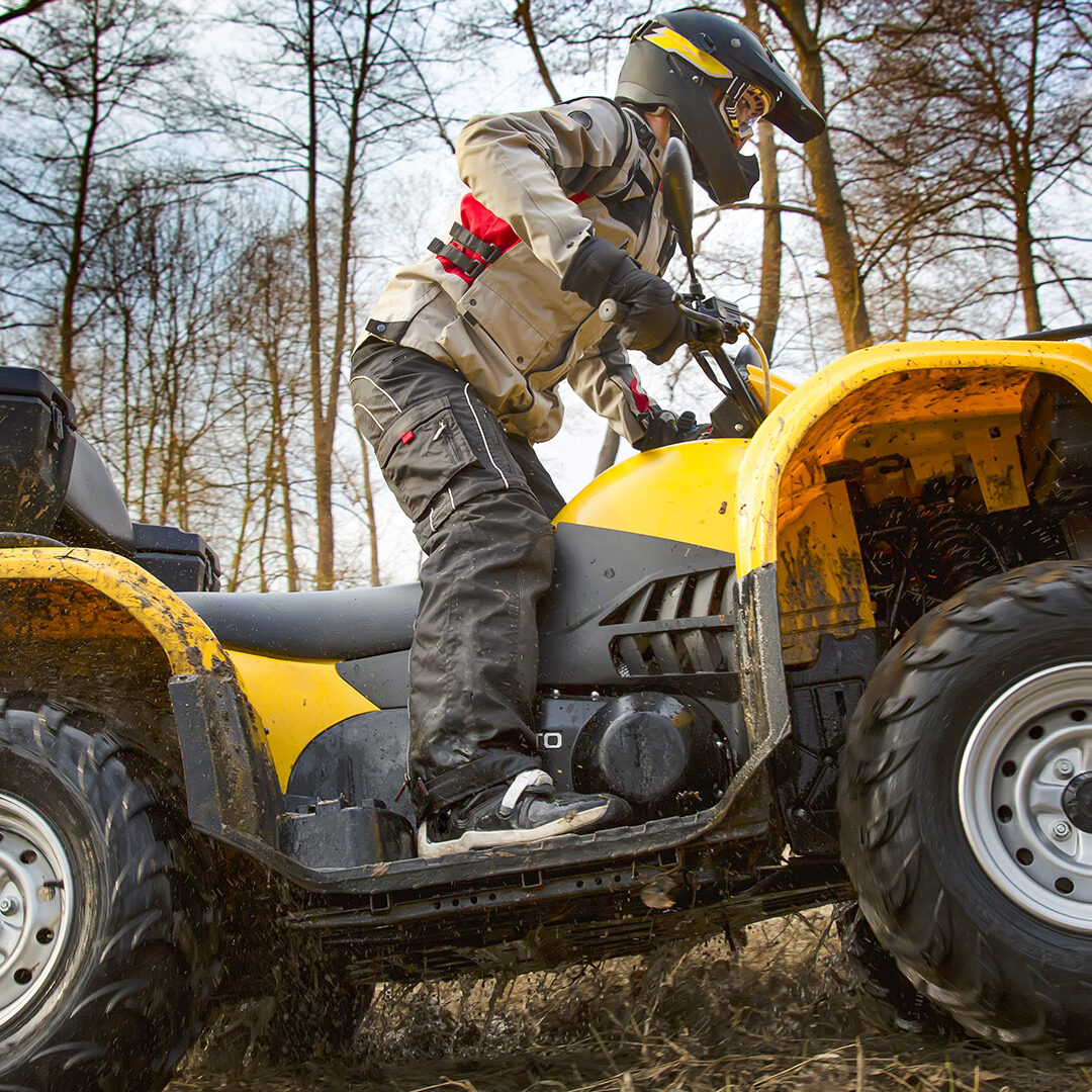 Horizontal motion portrait of a man in gray sport jacket and safety helmet and goggles driving mud-covered yellow ATV 4x4 quad bike with dirt spinning of the wheels.