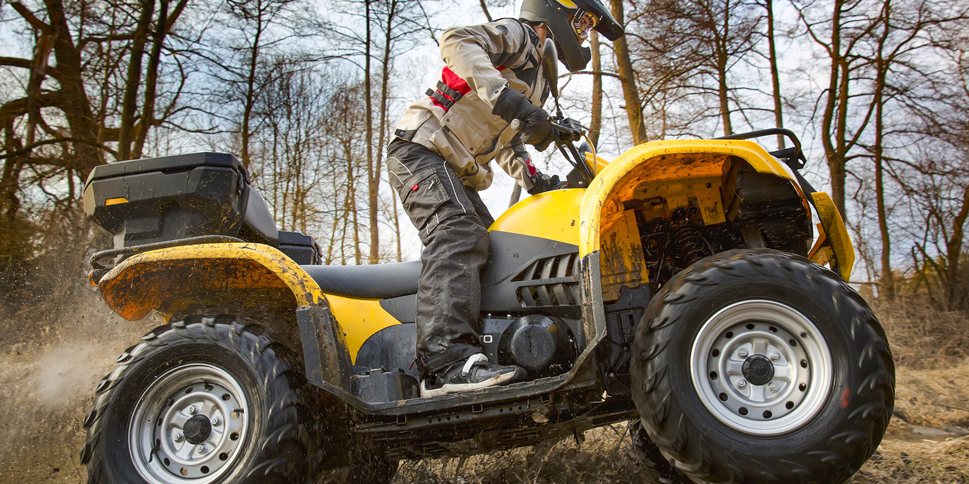 Dirt spinning of the ATV quad bike wheels Horizontal motion portrait of a man in gray sport jacket and safety helmet and goggles driving mud-covered yellow ATV 4x4 quad bike with dirt spinning of the wheels.