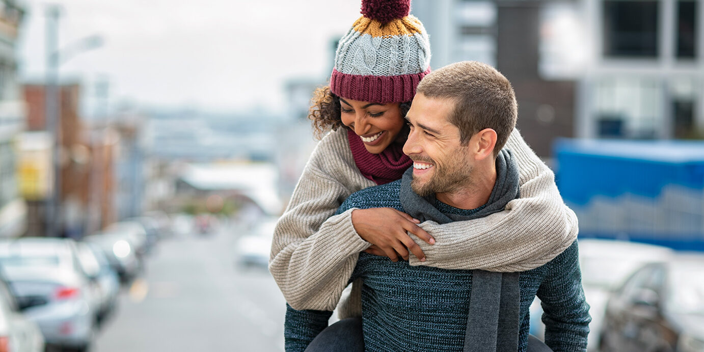 Smiling man giving piggyback ride to woman in the city. Young multiethnic couple in cold clothes walking in street and having fun. Cheerful girlfriend with wool cap and boyfriend in sweater enjoying winter together outdoor.