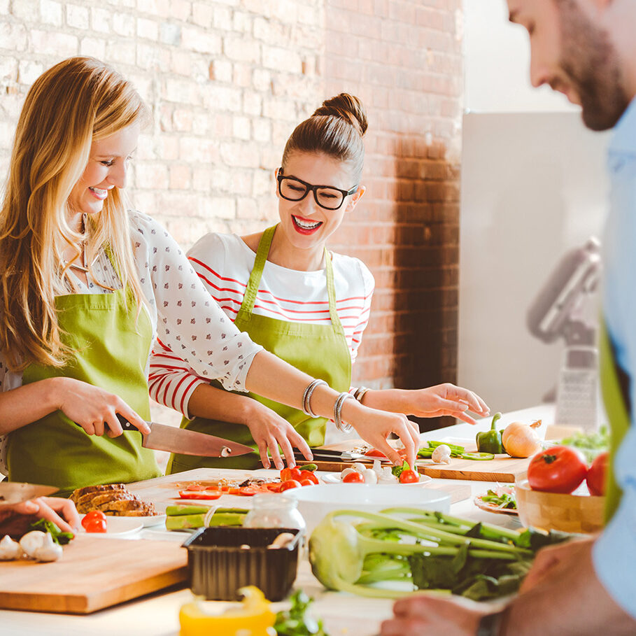 Group of people wearing aprons taking part in cooking class, preparing food, slicing vegetables, talking, laughing.