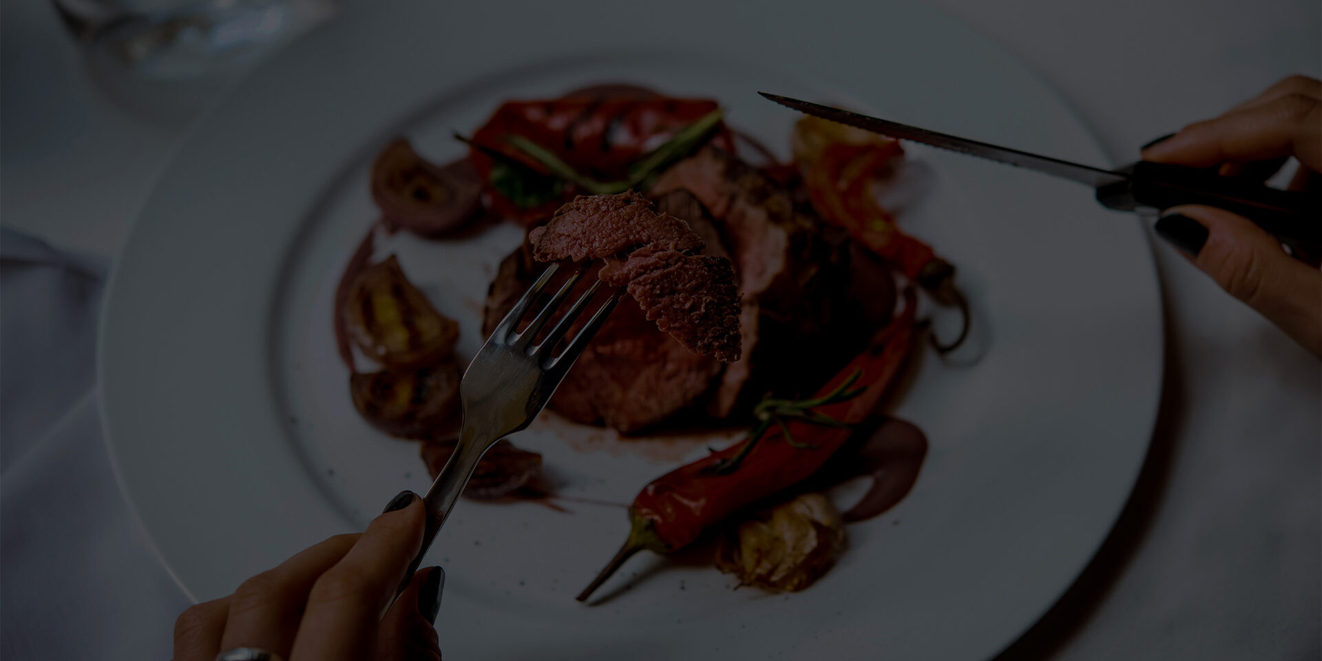 Person cuts beef steak with grilled vegetables served on white plate