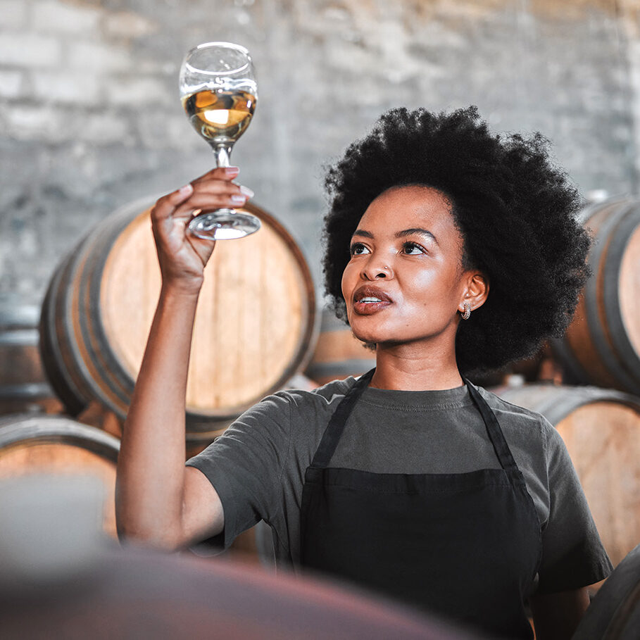 Black woman tasting wine at a winery, looking and checking the color and quality of the years produce. Young African American sommelier proud of the new addition, analyzing white wine in a cellar