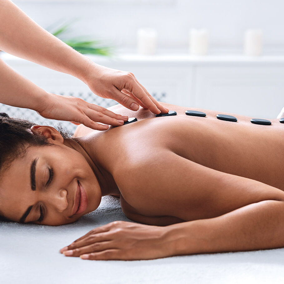 Therapist hands and young african woman enjoying hot stones massage, spa day concept