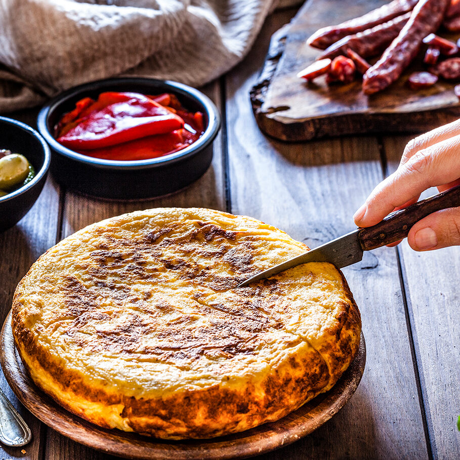 Spanish cuisine: Tapas. Rustic wooden table filled with traditional spanish tapas. At the center is a wooden plate with a spanish tortilla and a hand holding a kitchen knife is slicing the tortilla. A cutting board with spanish chorizo is at the top right. Bowls with olives, pimientos and peanuts complete the composition. Two bread slices are visible at the left. Predominant colors are yellow and brown. Low key DSRL studio photo taken with Canon EOS 5D Mk II and Canon EF 100mm f/2.8L Macro IS USM.