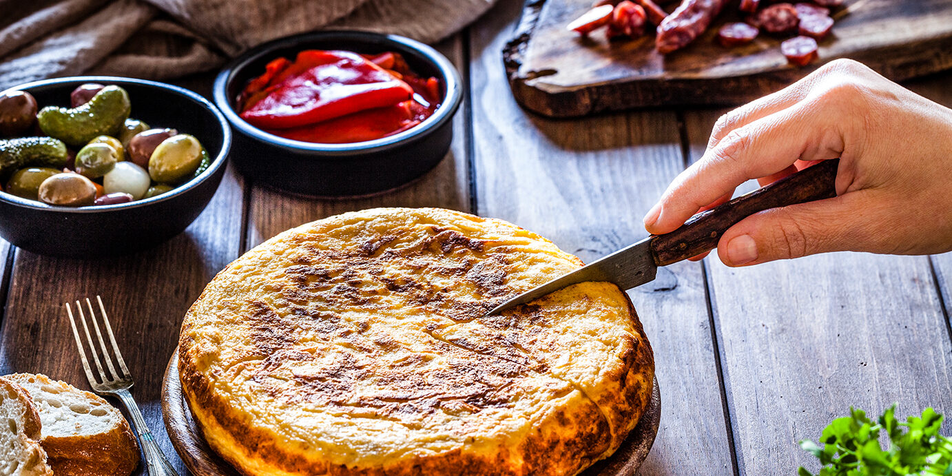 Tapas: Slicing spanish tortilla Spanish cuisine: Tapas. Rustic wooden table filled with traditional spanish tapas. At the center is a wooden plate with a spanish tortilla and a hand holding a kitchen knife is slicing the tortilla. A cutting board with spanish chorizo is at the top right. Bowls with olives, pimientos and peanuts complete the composition. Two bread slices are visible at the left. Predominant colors are yellow and brown. Low key DSRL studio photo taken with Canon EOS 5D Mk II and Canon EF 100mm f/2.8L Macro IS USM.
