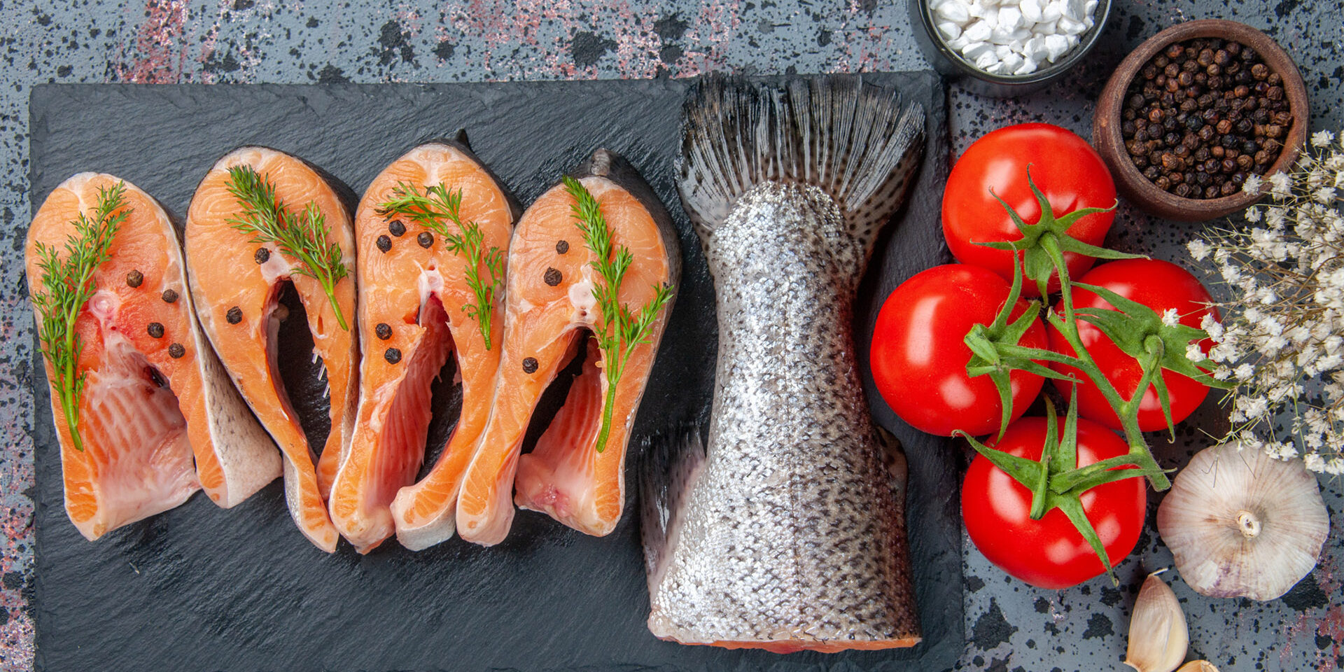 Top view of fresh raw fishes greens on dark color tray spices to Top view of fresh raw fishes greens on dark color tray spices tomatoes with stems garlics flowers on blue black mix colors background