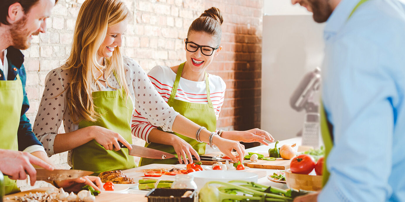 Four people taking part in cooking class Group of people wearing aprons taking part in cooking class, preparing food, slicing vegetables, talking, laughing.
