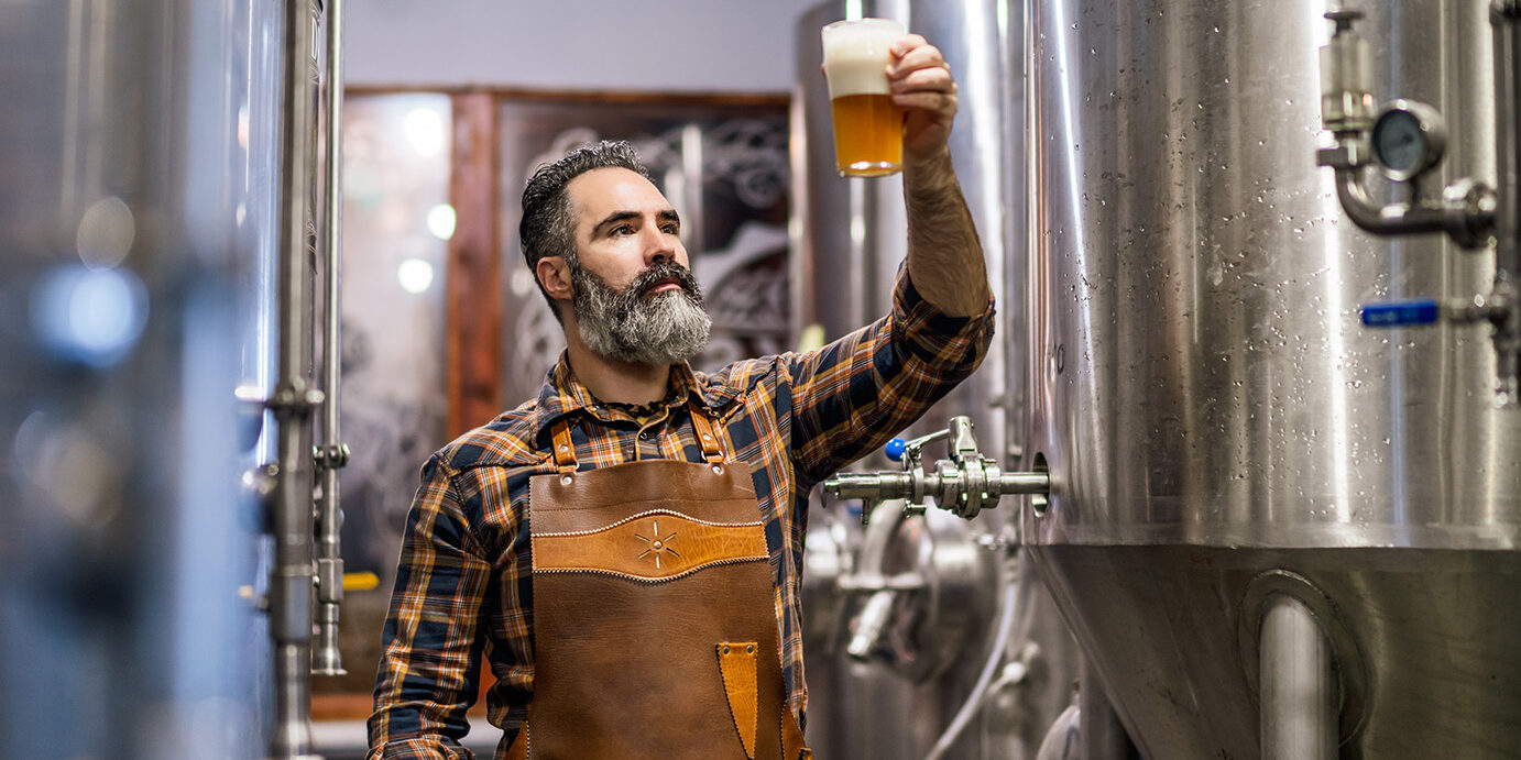 Craft beer production Bearded brewery master holding glass of beer and evaluating its visual characteristics. Small family business, production of craft beer.