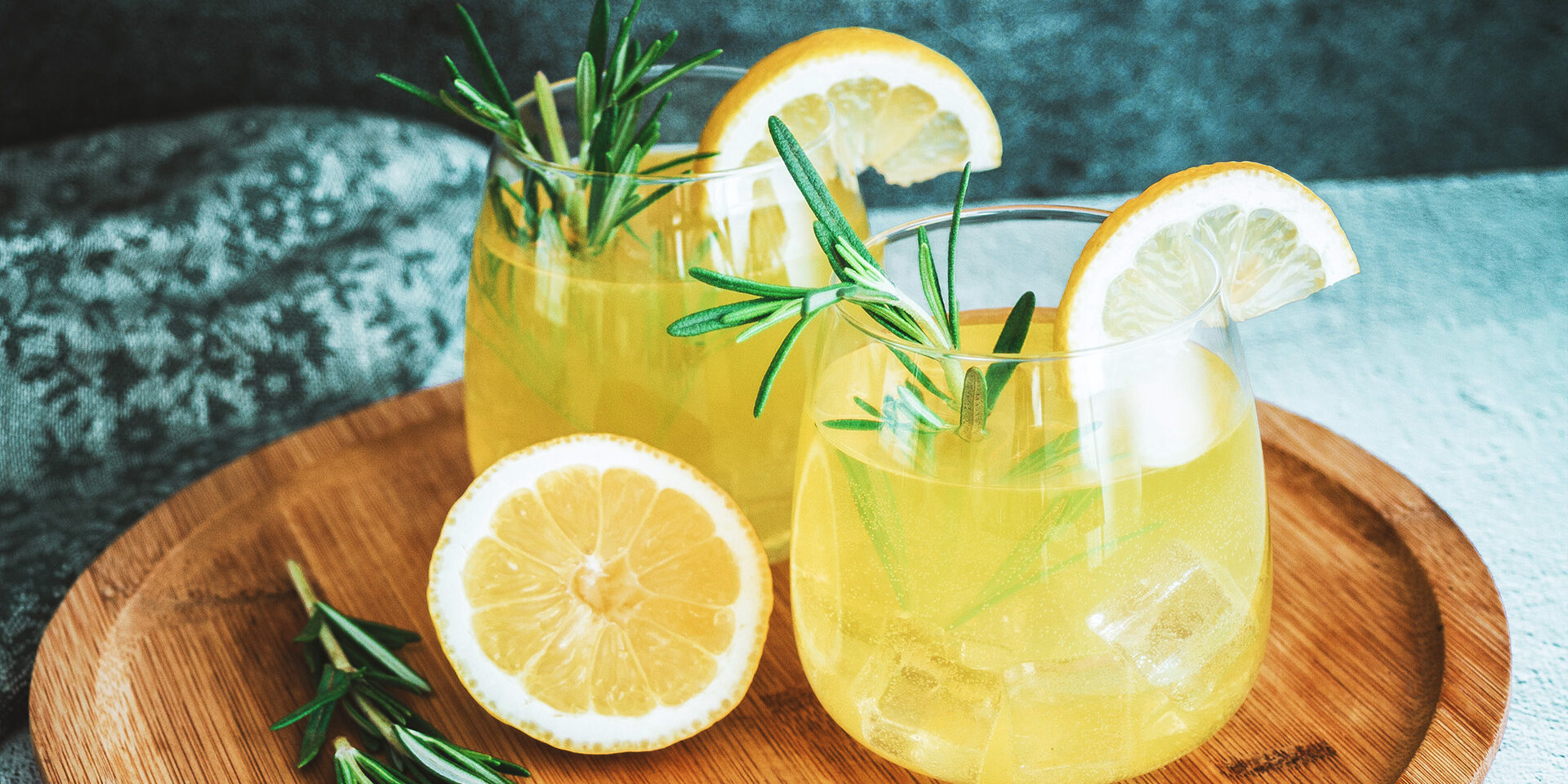 Two glasses with cold yellow cocktail, lemonade, sliced lemon and rosemary plant on the wooden plate. Drink close up still life on the white table and concrete grey wall background. Horizontal photo for bar, menu content.