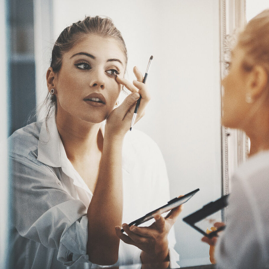 Closeup of attractive mid 20's blond woman putting on some makeup in front of large bathroom mirror. Shot from behind, focus is on her reflection. Softly toned and desaturated.