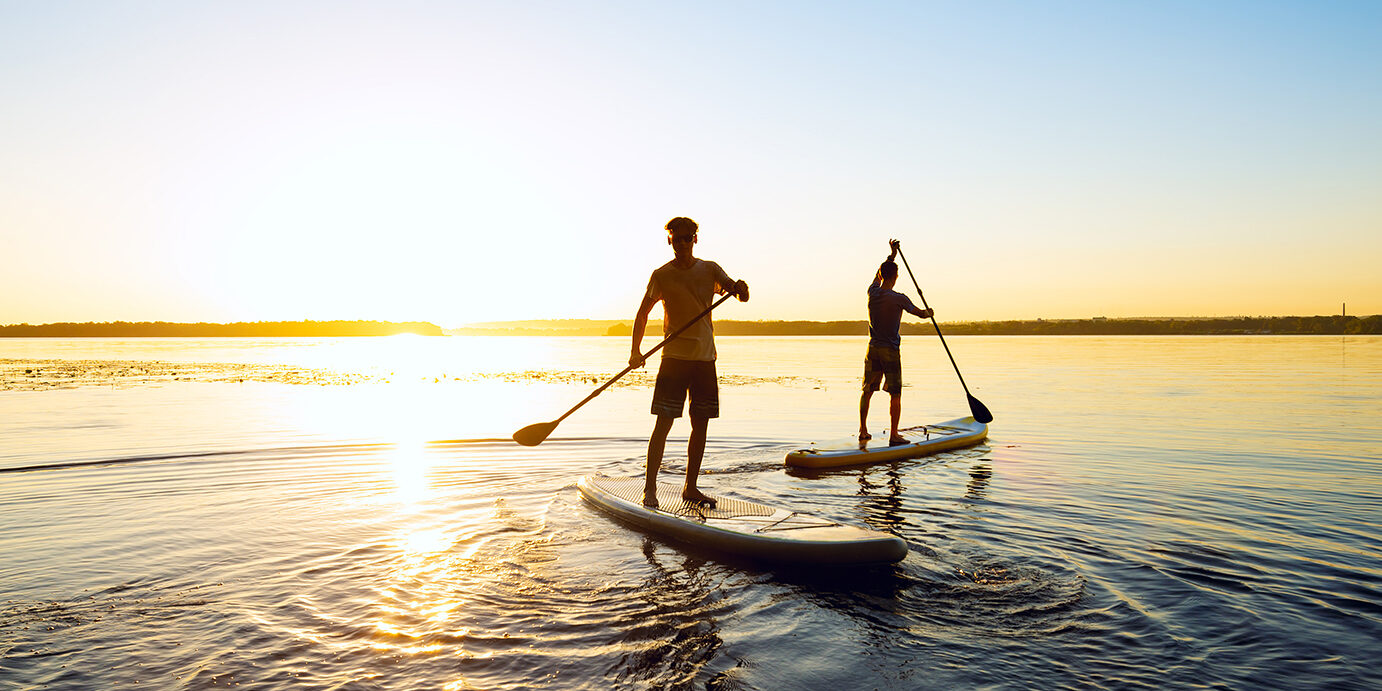 Men, friends are paddling on a SUP boards on a large river during sunrise. Stand up paddle boarding - awesome active recreation in nature. Backlight.