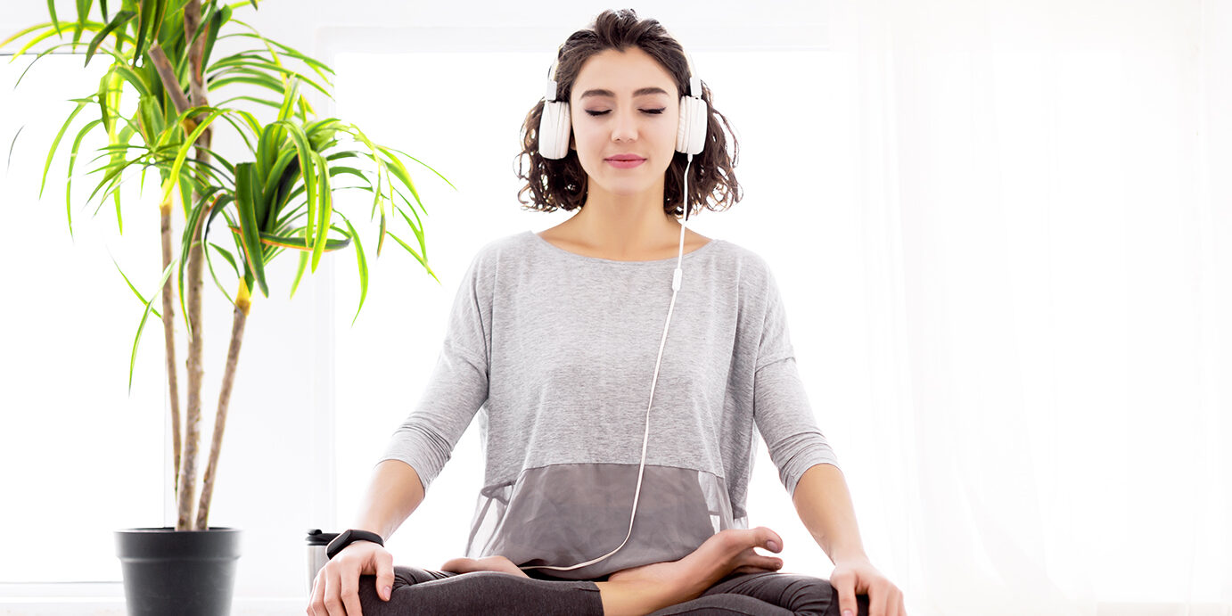 Modern woman with headphones sitting in yoga lotus posture Slender female folded crossed legs in lotus position. Young athletic woman in fitness tracker and headphones is practicing yoga, meditating, relaxing while sitting in padma asana at home.