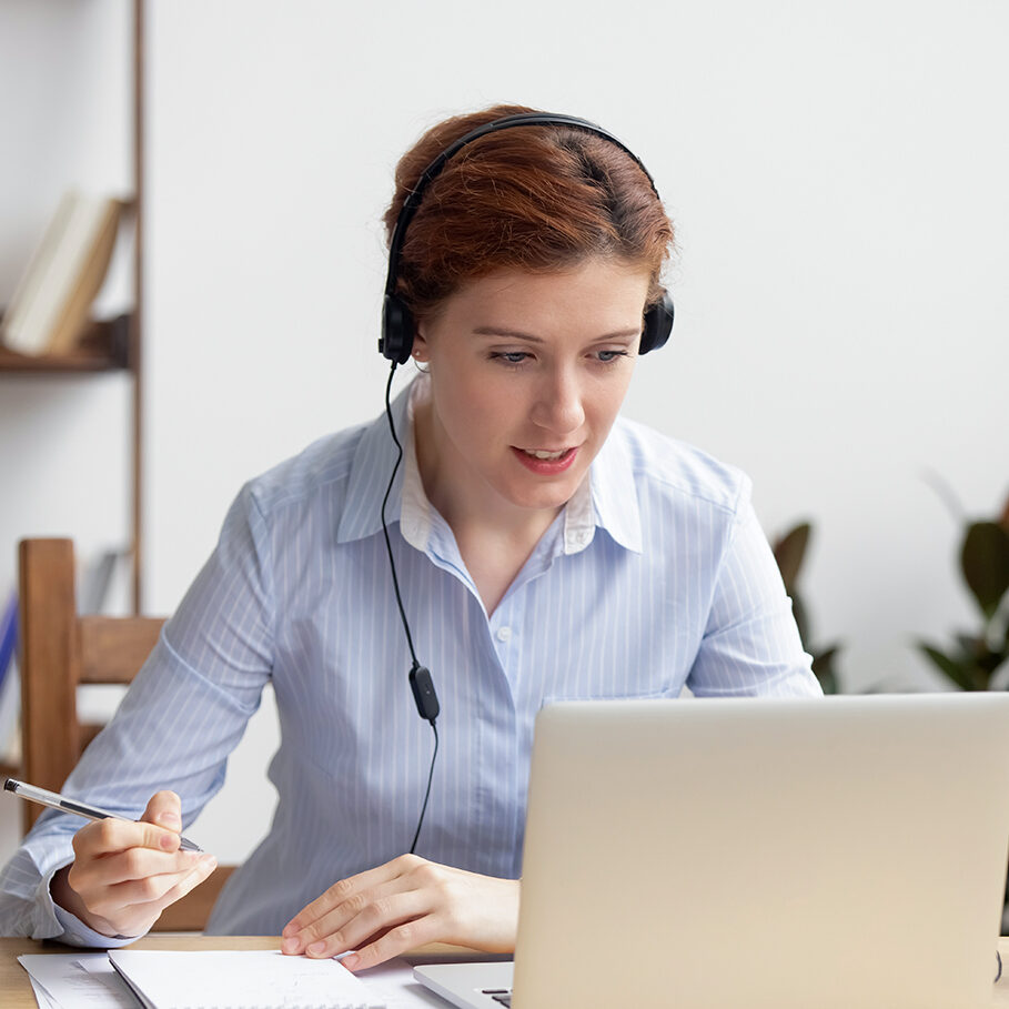 Businesswoman wearing headphones watching video webinar making conference online call writing notes talking on skype, focused woman study online looking at laptop listening translating lecture course