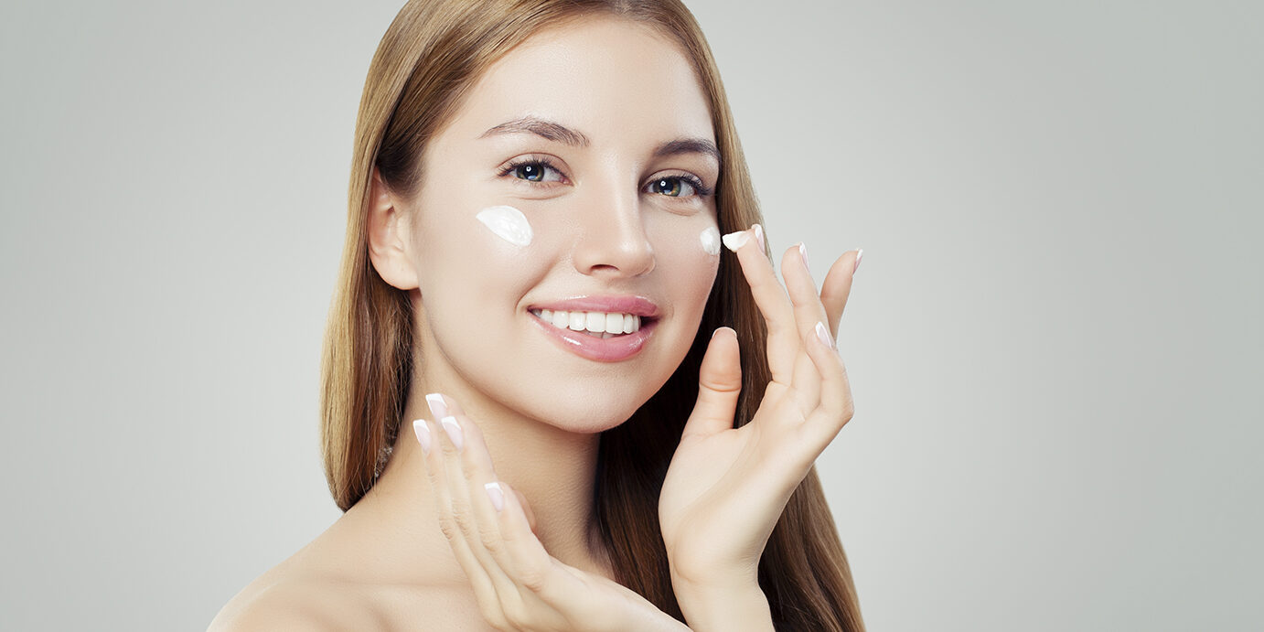Happy young woman applying cream on her face. Skin care, beauty and facial treatment concept Happy young woman applying cream on her face. Skin care, beauty and facial treatment concept