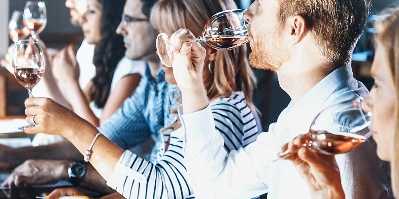 Wine tasting. Closeup of group people tasting different types of wines and discussing about visual appearance, smell, flavor and taste. There are three men and three women, seated side by side in a tasting room. Focus on the closest person to the camera.