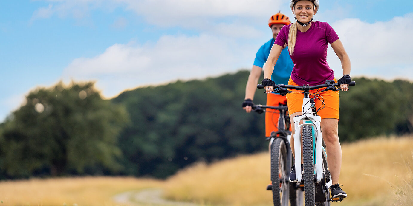 happy smiling couple cycling down gravel road in rural landscape with elctric mountain bikes in summer, focus on woman, place for text