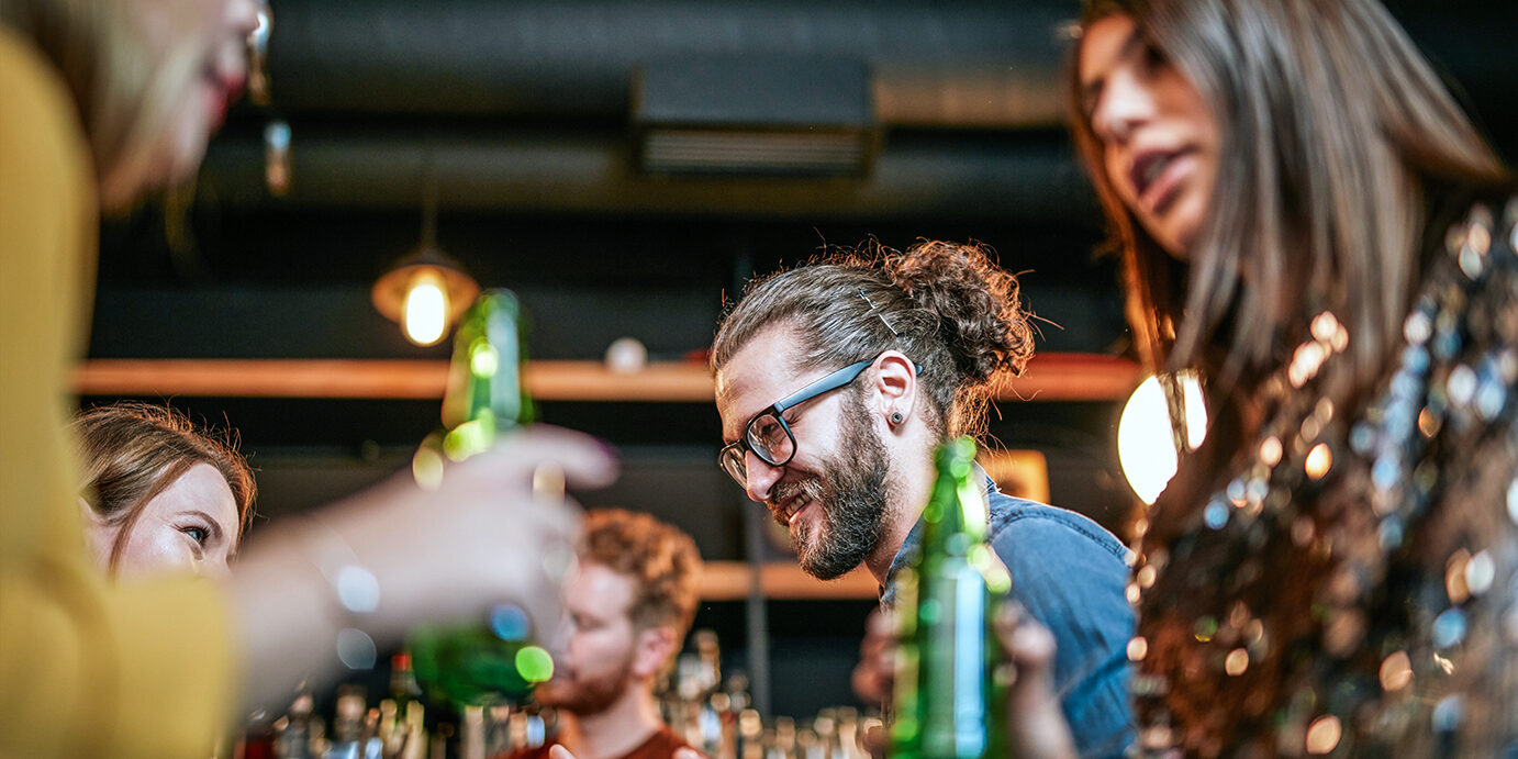 A small group of best friends standing at a pub, drinking beer, chatting and having fun. Focus on bartender wiping drinking glass. Nightlife.