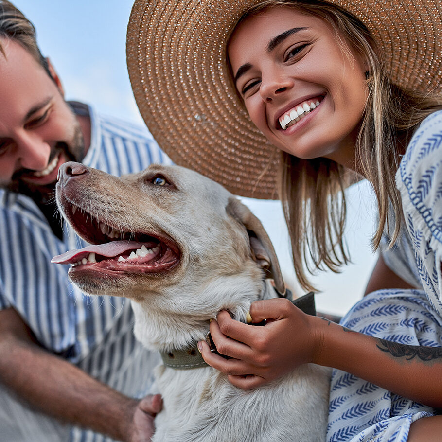 A cute woman in a dress and a straw hat and a handsome man in a striped shirt with their labrador dog are having fun on the seashore.