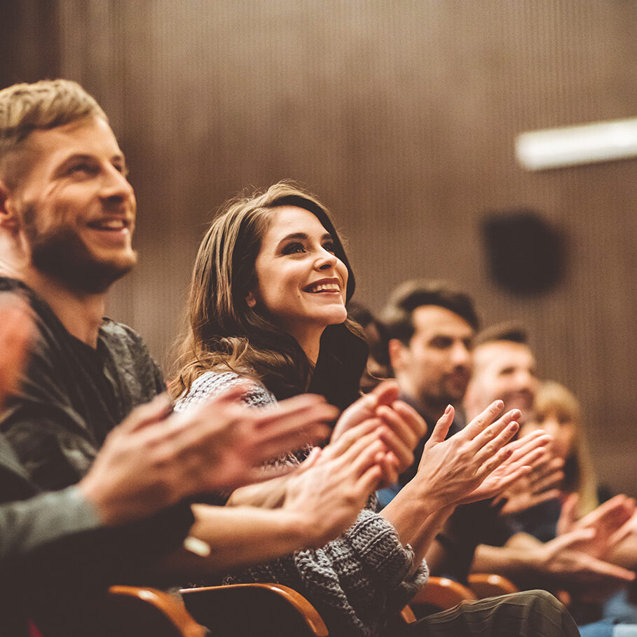 Group of smiling people clapping hands in the theater, focus on audience. Dark tone.