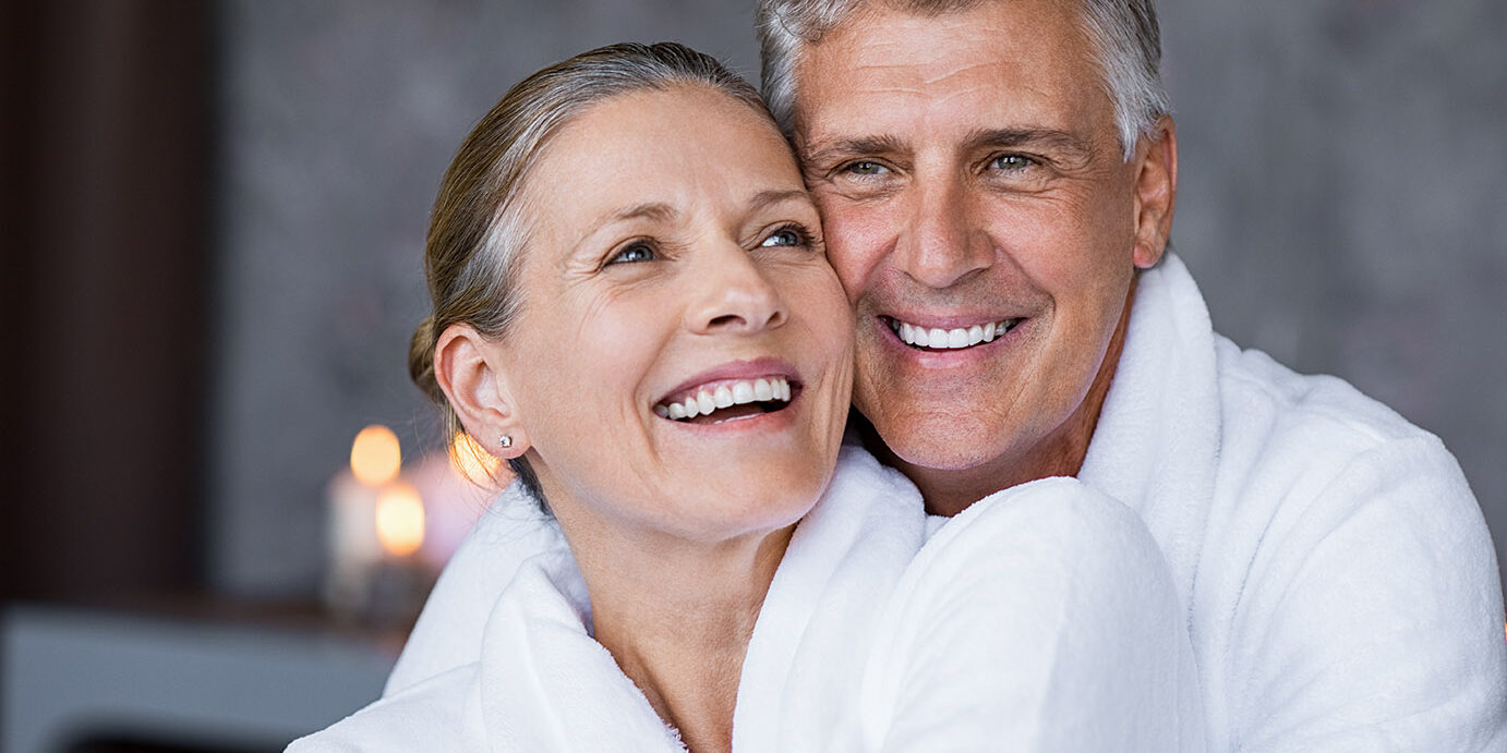 Smiling husband embracing cheerful wife from behind at spa. Laughing mature couple enjoying a romantic hug at wellness center after massage. Senior man and woman in white in bathrobe relaxing at spa.