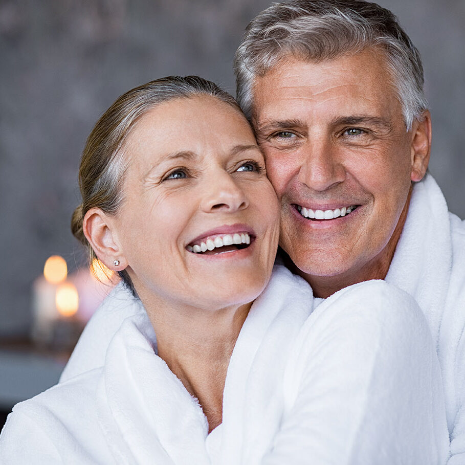 Smiling husband embracing cheerful wife from behind at spa. Laughing mature couple enjoying a romantic hug at wellness center after massage. Senior man and woman in white in bathrobe relaxing at spa.