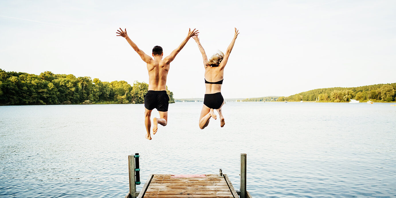 Two friends jumping with their arms in the air off the end of a jetty at a lake together.