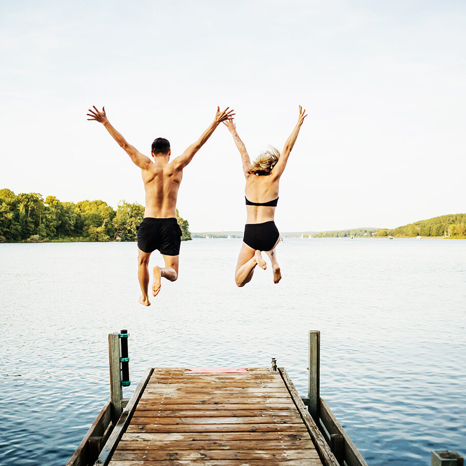 Two friends jumping with their arms in the air off the end of a jetty at a lake together.