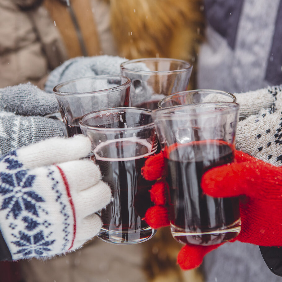 Close up of hands holding cups with hot wine. Friends cheering with glasses. Wearing warm knitted gloves.