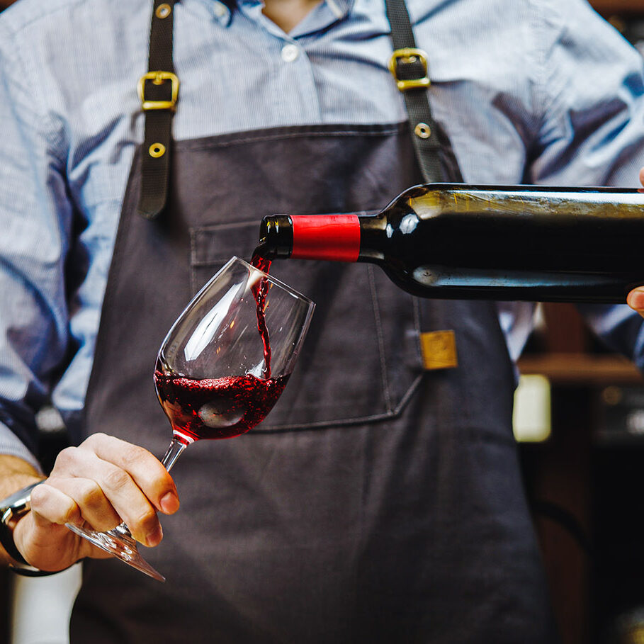 Male sommelier pouring red wine into long-stemmed wineglasses. Waiter with bottle of alcohol beverage. Bartender at bar counter pour elite drink into long-stemmed glass