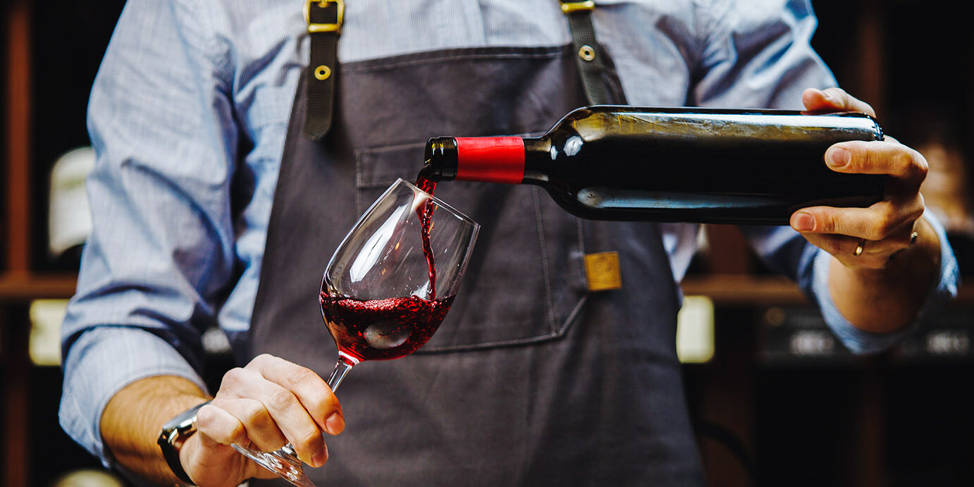 Male sommelier pouring red wine into long-stemmed wineglasses. Male sommelier pouring red wine into long-stemmed wineglasses. Waiter with bottle of alcohol beverage. Bartender at bar counter pour elite drink into long-stemmed glass