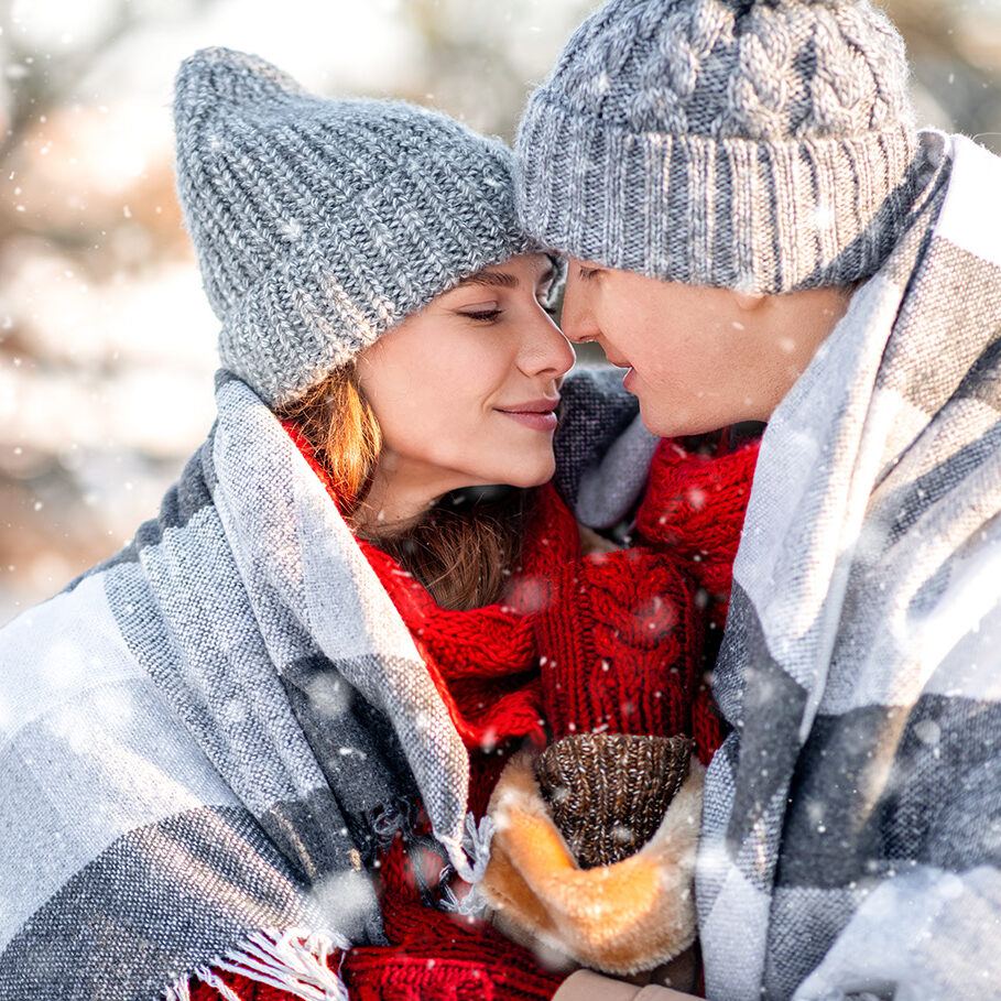 Closeup portrait of romantic couple enjoying snowy winter day, beautiful young man and woman in winter outfits covered in blanket cuddling, embracing, touching each other with noses